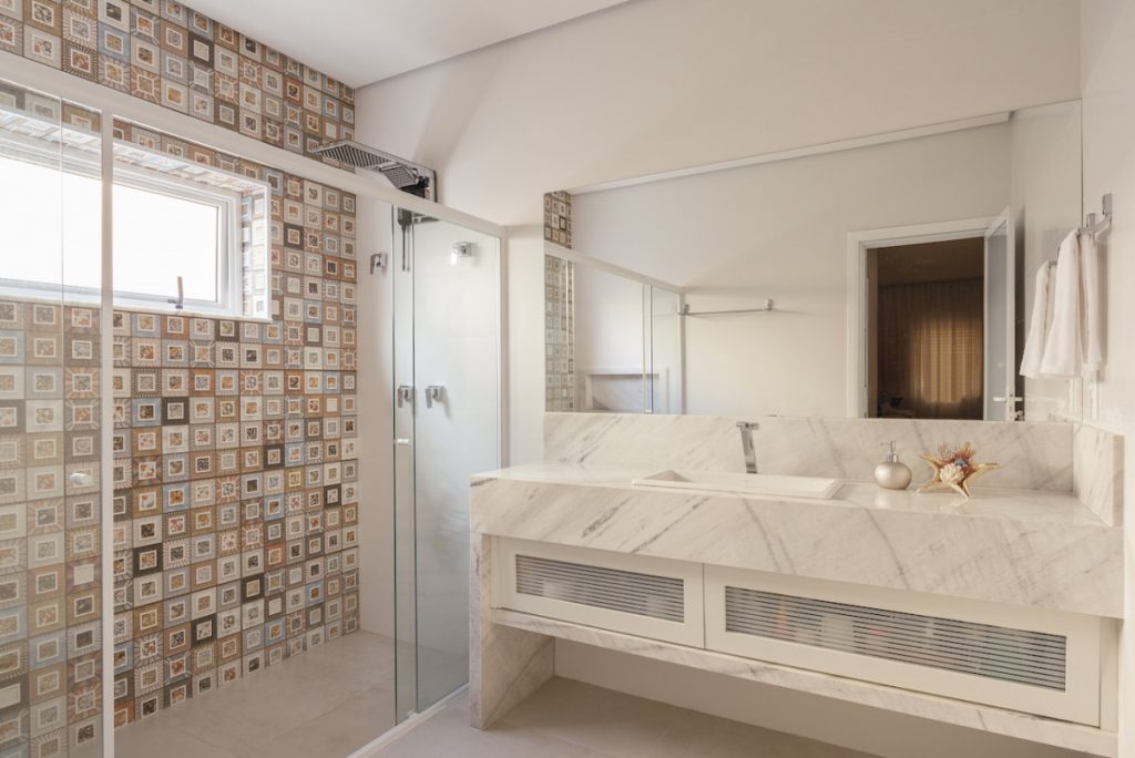 Banheiro de uma casa vazio, com mármore branco, piso bege, box de vidro e chuveiro.