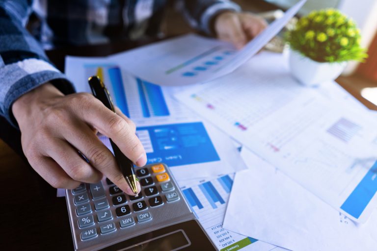 Mão de um contador com caneta, papéis e calculadora na mesa, fazendo os cálculos da declaração do imposto de renda