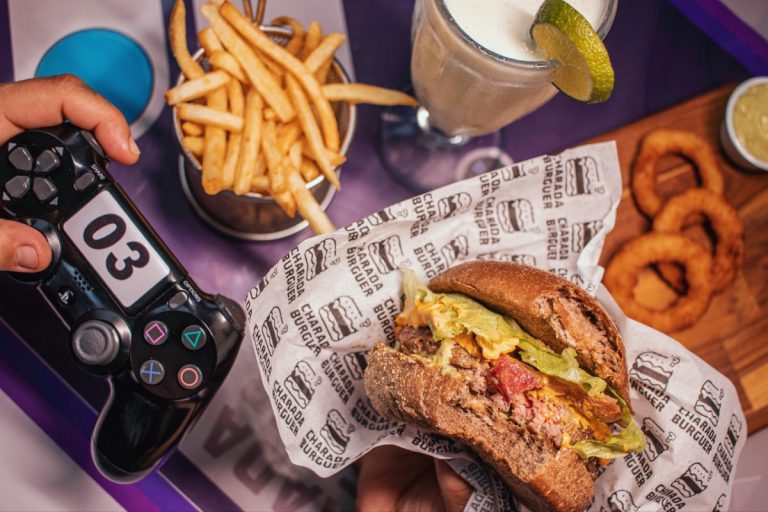 Mesa com hamburguer, batata frita, rodelas de cebola e um clinte com controle de playstation na mão