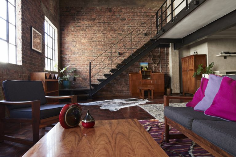 Na imagem, vemos uma sala de um apartamento com chão de madeira, paredes de tijolos e com decoração moderna
