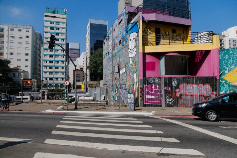 Na imagem vemos imóveis em fase de demolição ao lado de um estacionamento em avenida movimentada em São Paulo