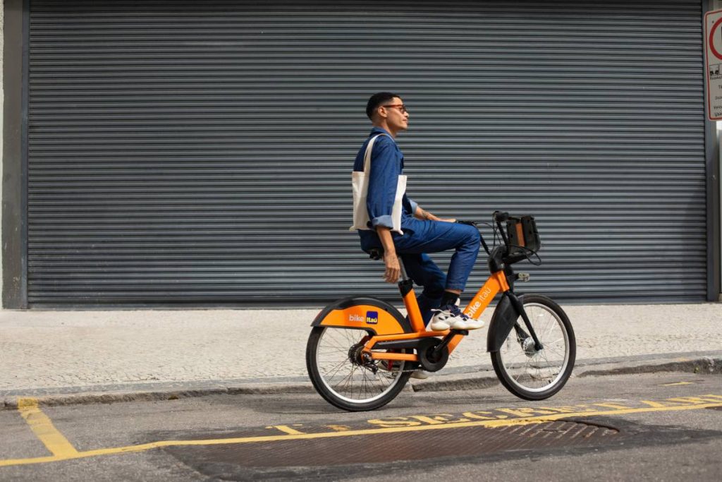 Homem andando com uma bicicleta laranja da Tembici, startup para cidades inteligentes com foco em transformar a circulação de pessoas